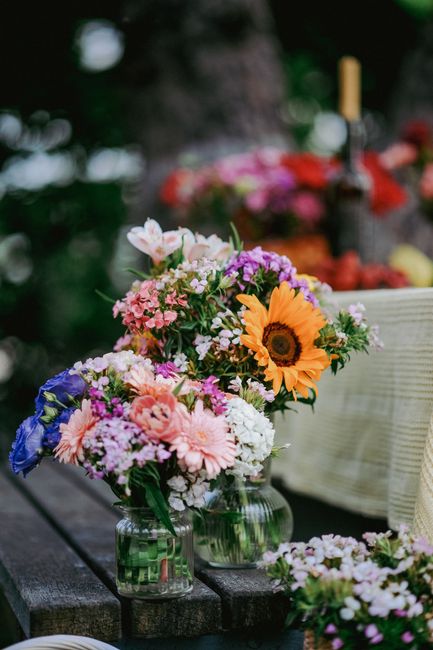 A collection of fresh floral arrangements featuring a variety of colorful flowers in small glass jars and containers, placed on a dark wooden outdoor table. The bouquets include a prominent sunflower