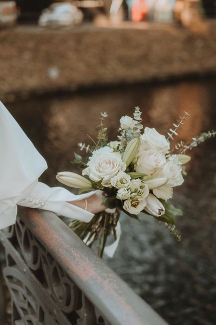 A bridal bouquet featuring white roses, lilies, and small white spray flowers arranged with green foliage, including eucalyptus and other textured leaves. The bouquet is wrapped in white satin ribbon