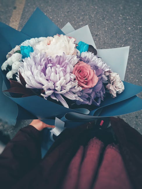 A person holding a floral bouquet wrapped in blue and white paper, featuring a variety of fresh flowers including white chrysanthemums, a pink spray rose, and purple carnations, arranged with green st