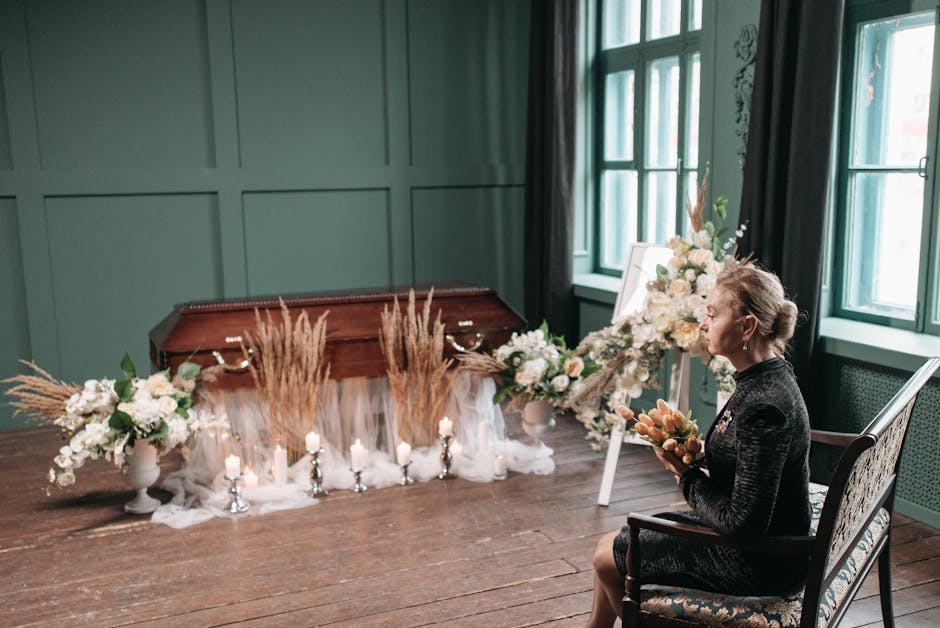 A woman seated indoors on a vintage armchair holds a bouquet of soft peach and pink flowers. In front of her, two large floral arrangements sit on the dark wooden floor, featuring white roses, peonies