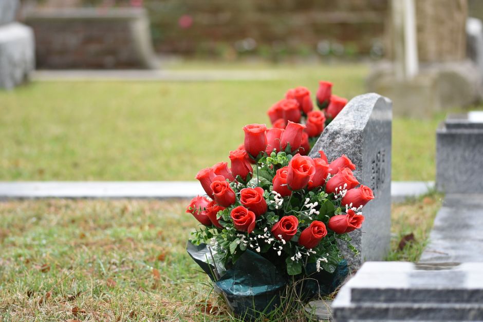 A floral arrangement of fresh red roses with tightly closed petals sits against a grey gravestone in a cemetery. The bouquet is wrapped in dark green floral plastic and includes small white baby's bre