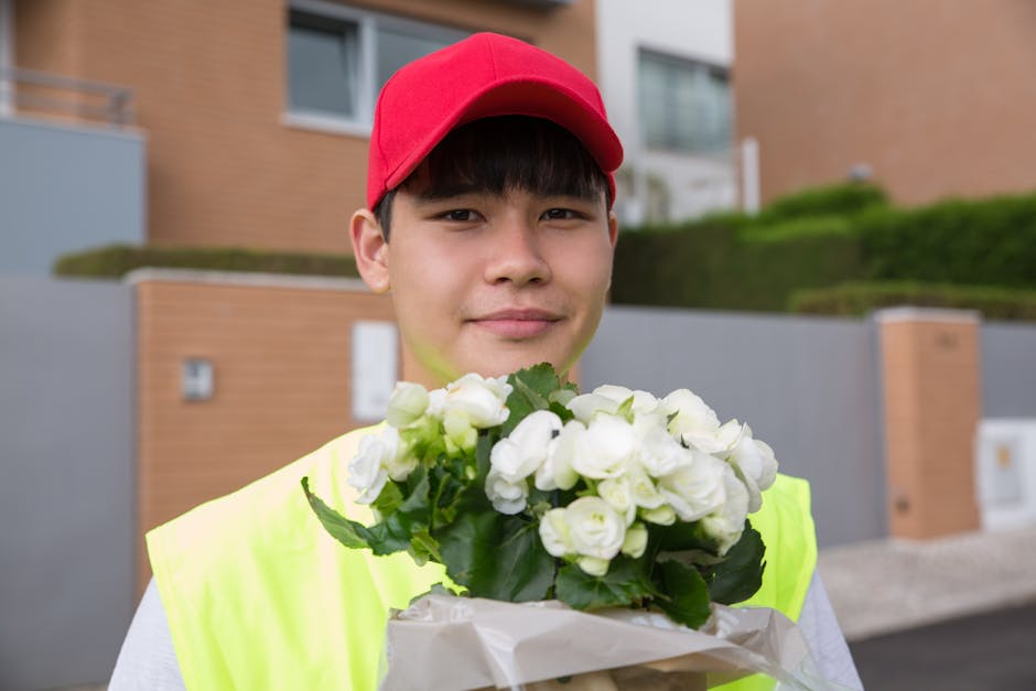 A young delivery person wearing a red cap and a high-visibility yellow vest is standing outdoors in a residential area with modern, brick and concrete buildings in the background. They are holding a b