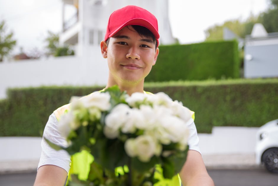 A delivery person in a red cap and high-visibility vest smiling while holding a bouquet of white roses and green foliage, with a blurred background of residential buildings, hedges, and a parked white