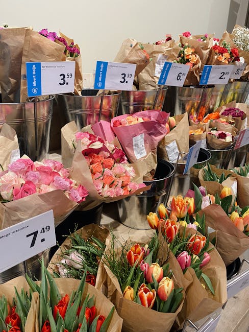 A display of freshly arranged floral bouquets featuring pink roses, orange and yellow tulips, and mixed pink and red flowers wrapped in brown craft paper. The flowers are organized in metallic vases w