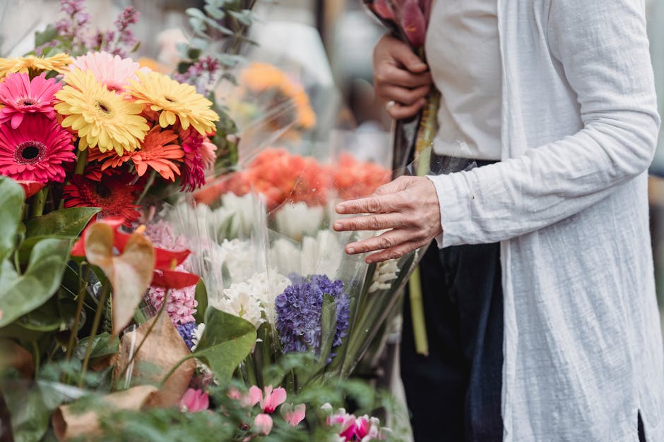 A person wearing a light grey long-sleeved coat is examining a large bouquet of fresh flowers at a flower market, with their right hand gently touching the flowers. The bouquet includes vibrant pink, 