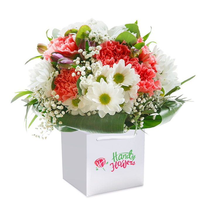 Arranged with a craftsman's precision and deep local pride, this vibrant Cubitt Town bouquet displays a harmonious interplay of colour and texture inside a flawless square white gift bag. At its core, plush coral and striking red carnations-richly ruffled in layers-invite a sense of indulgent beauty, each petal seemingly brushed with sunlight. They are joined by crisp white daisies, their lively chartreuse eyes sparkling with warmth, reminiscent of cheerful scenes along Stebondale Street. Throughout the bouquet, delicate baby's breath threads gentle clouds of white between the larger blooms, softening the composition and imparting a natural, effortless grace. Awaiting discovery, a few closed buds edged with purple and pink add a whisper of mystery and depth, hinting at stories yet to bloom. Embracing the arrangement, a lush wreath of deep green, broad-leaved foliage provides structure and polish, echoing the calm verdancy lining the riverside walks. The gift bag, enhanced with sturdy ribbon handles for smooth delivery anywhere in Cubitt Town, features discreet branding that assures quality without distraction. The entire image, professionally lit to showcase each detail from petal to leaf, resonates with freshness and careful presentation. Perfect for celebrating birthdays, tokens of appreciation, or milestone moments, this bouquet serves as a vibrant expression of floral artistry delivered from the heart of the community.
