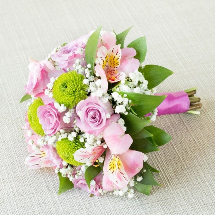 A hand-tied floral masterpiece designed for weddings, this bouquet is displayed diagonally atop a pale beige textile, calling to mind the simple elegance of Cubitt Town's riverside interiors. Blush and rose-pink blooms unfurl with gentle fragrance, their velvety petals forming delicate spirals that anchor the bouquet with romantic appeal. Peppering the arrangement are vivacious lime-green button chrysanthemums-tiny spheres of freshness and contrast to the pastel tones-while soft pink alstroemeria open wide, revealing inner petals speckled with yellow and burgundy, reminiscent of flora glimpsed by Canary Wharf's gardens. Ethereal sprays of white baby's breath drift through the arrangement, lending a light, airy aura that ties the elements together. Glossy deep green leaves meander through the bouquet, enhancing its sense of depth and highlighting the floral colours. At the foundation, stems are expertly bundled and secured with a sleek fuchsia ribbon, tied firmly to ensure a modern presentation with the natural stems exposed beneath-a nod to authentic florist technique. Bathed in gentle daylight, this bouquet encapsulates themes of love, celebration, and freshness, suitable for Cubitt Town's treasured events or as an uplifting local gift, and crafted with skill by the team at Cubitt Town Flowers.