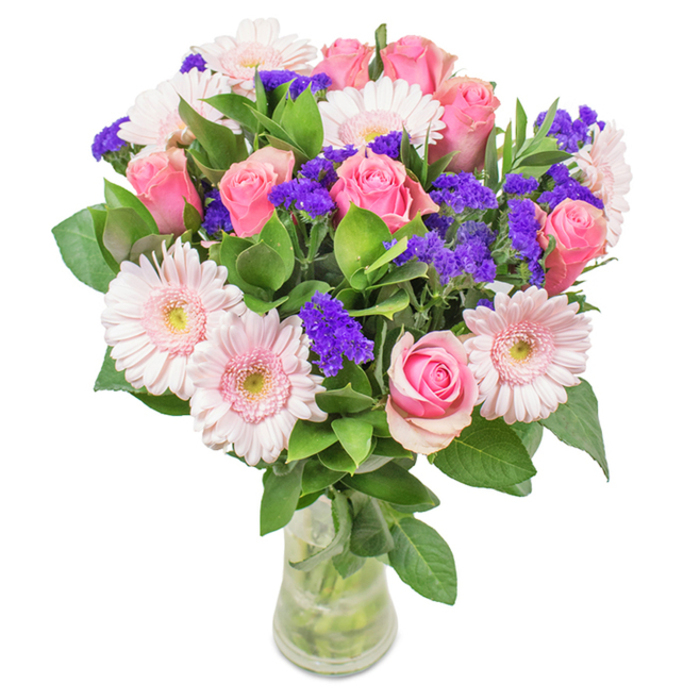This sophisticated floral arrangement, crafted with care by Cubitt Town Flowers, sits gracefully in a clear glass vase against a brilliant white background. The ensemble showcases around seven to nine pink roses at varying stages of bloom, their petals coiled in classic spirals or just gently opening, celebrating natural form and subtle gradients of pink. Framing the roses, five to seven pale pink gerbera daisies-recognisable by their full, round faces and nearly white centres-add a layer of textured softness. Sprays of vivid purple statice are woven throughout, their colour echoing local flower market vibrancy while offering visual and tactile contrast. A lush halo of deep green foliage grounds the bouquet, with each stem's freshness confirmed by clear water visible through the glass. The arrangement, available for same-day delivery in Cubitt Town, is equally suitable for birthdays, anniversaries, or as a thoughtful decorative piece for riverside apartments, carrying with it an enduring impression of joy, affection, and attentive Cubitt Town floristry.